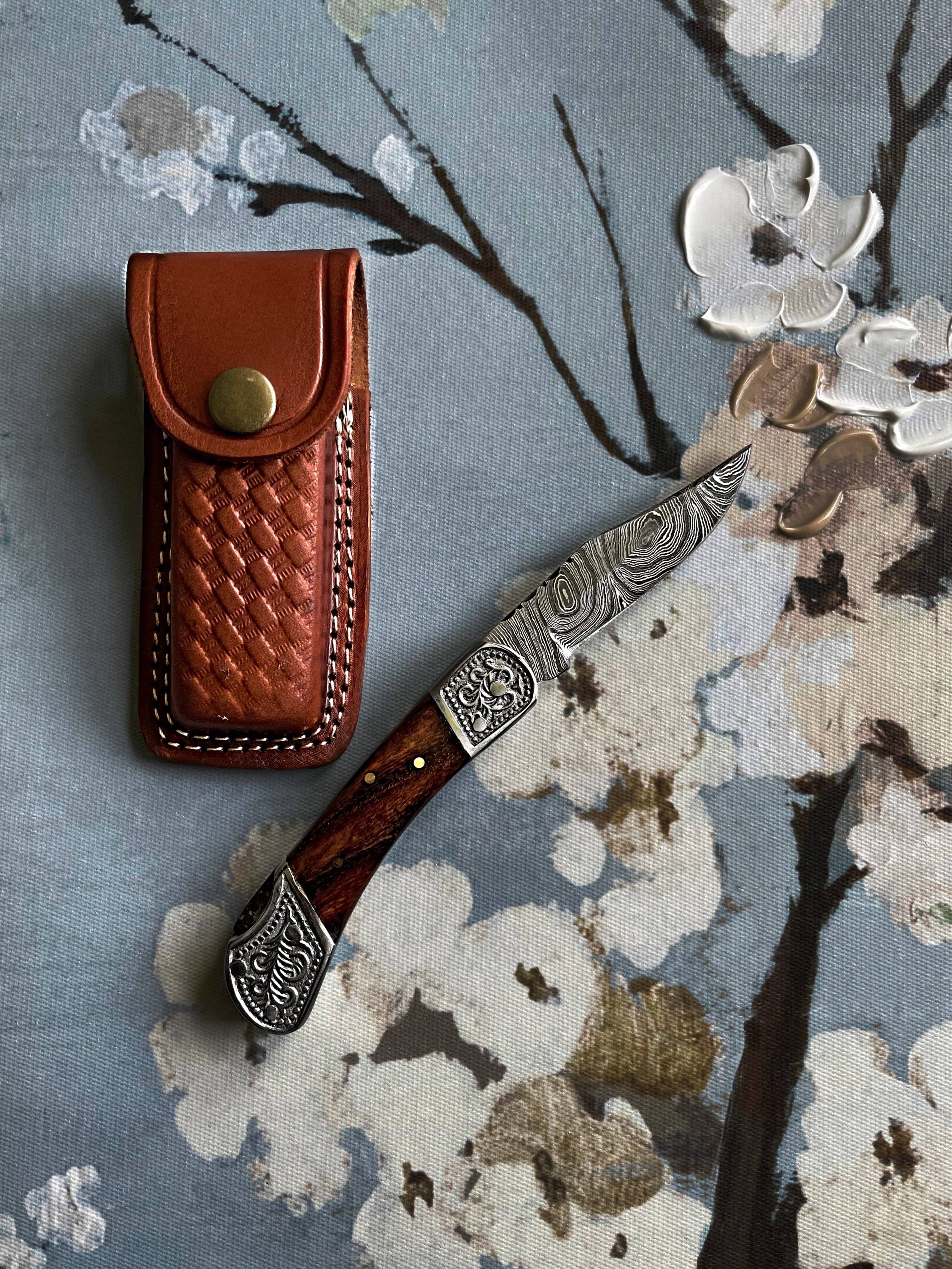 The Pocket Knife Walnut Wood 6.5'' Damascus Steel, featuring an engraved blade and walnut handle, is displayed beside a brown leather sheath on a floral-patterned canvas background.