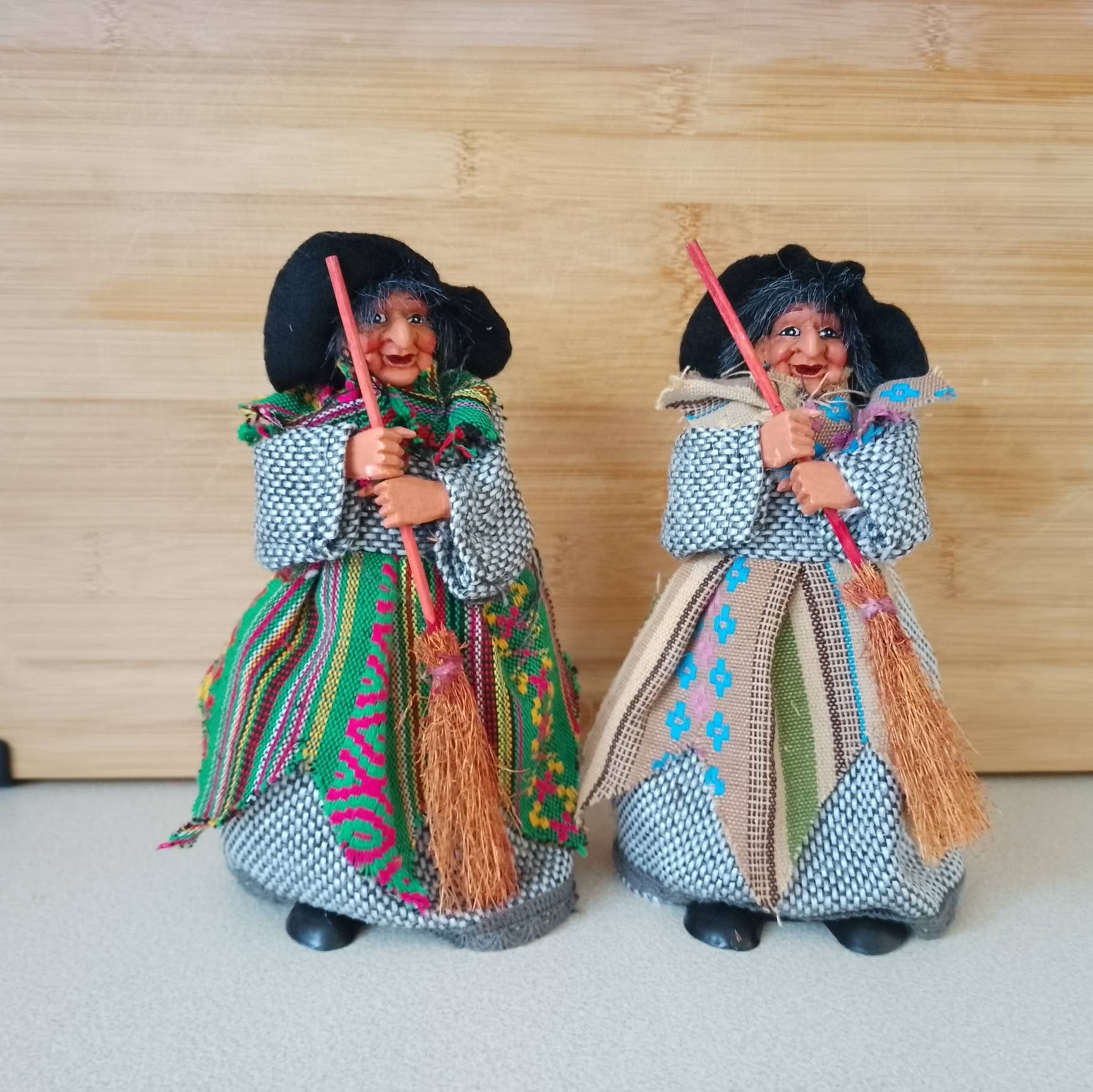 Two Yasmina Green Kitchen Witch figurines, each 7" tall and holding brooms, stand side by side on a light surface with a wooden background.