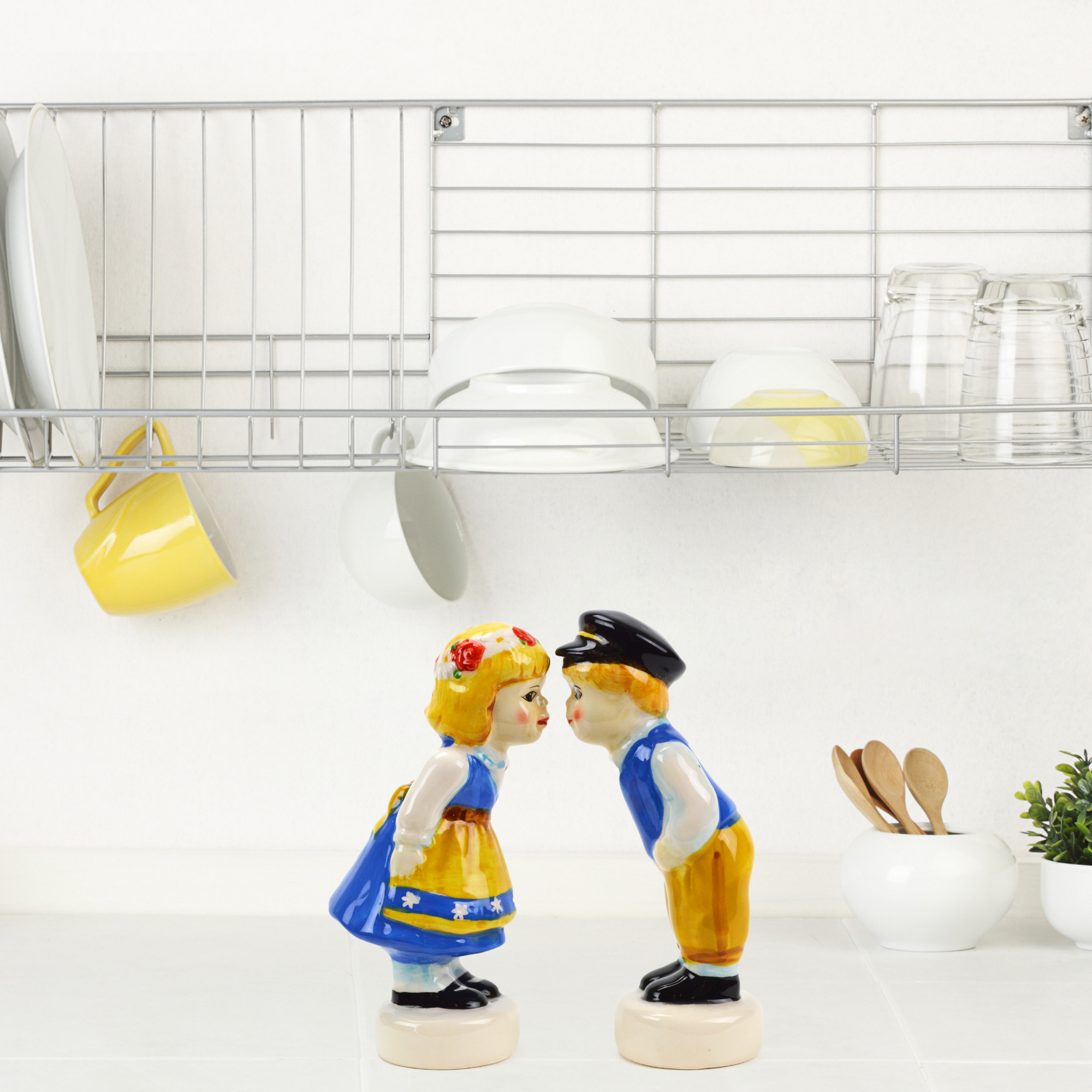 Two ceramic figurines from the Vintage Salt & Pepper Shakers Swedish Standing Couple set face each other on a kitchen counter. Above them, a dish rack holds plates, cups, and glasses. Nearby, a small potted plant adds a touch of greenery to the delightful Swedish kitchen decor.