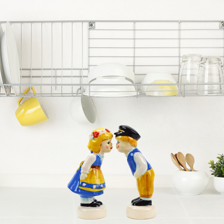 On the counter, two ceramic figurines, a boy and a girl, evoke vintage kitchen charm as they face each other beneath a dish rack with plates, cups, and glasses. These delightful pieces could easily be mistaken for Swedish Standing Couple Vintage Salt & Pepper Shakers.