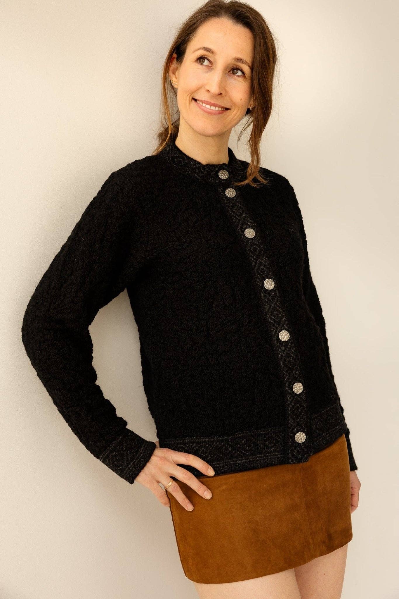 A woman stands against a light background, smiling and looking to the side in the Bryggen Women's Norwegian Cardigan (White), featuring silver buttons and 100% wool, paired with a brown suede skirt.