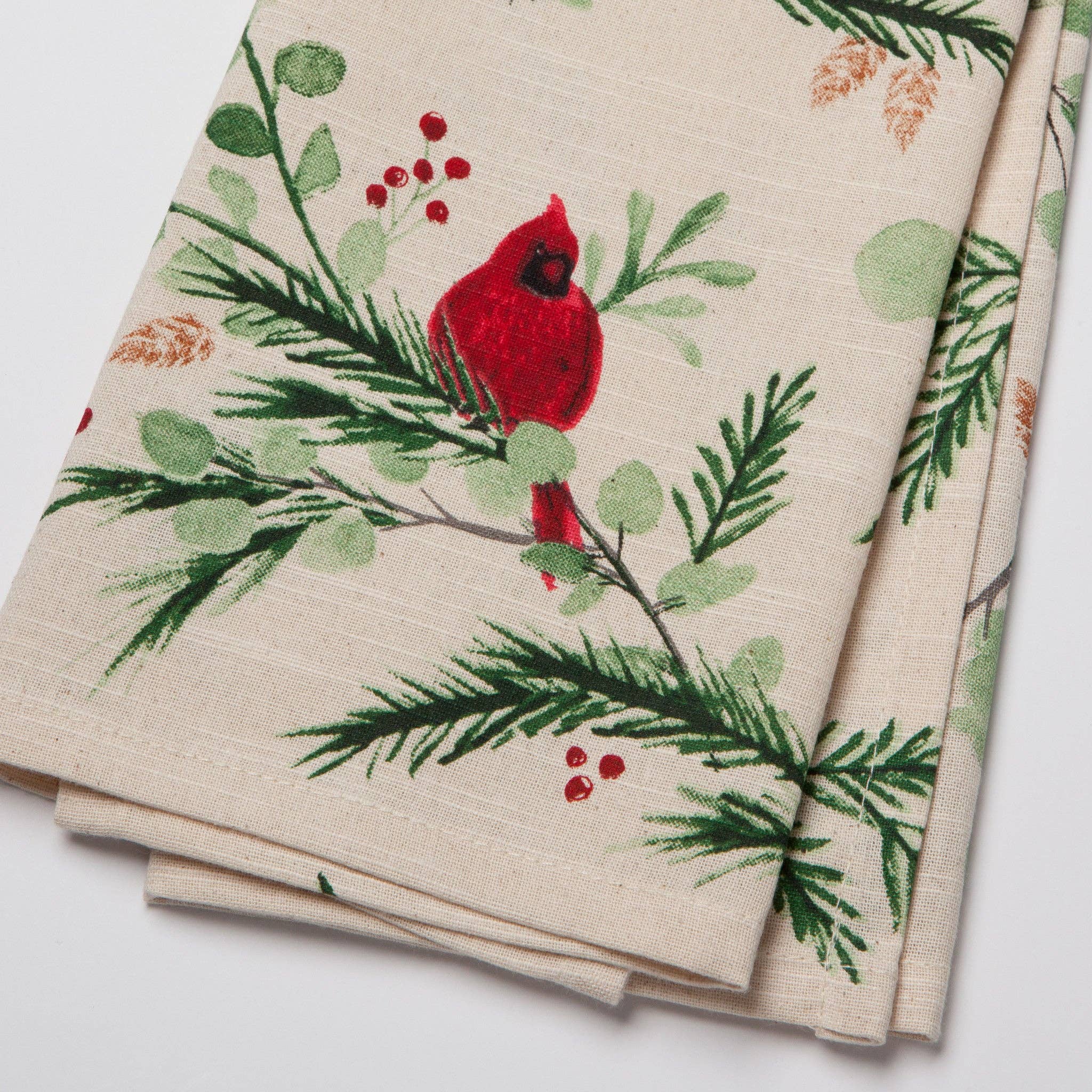 The Tea Towel: Forest Birds Christmas Dishtowel boasts a beige fabric with a rustic charm, highlighting a red cardinal and green foliage design, including pine branches and red berries, evoking the essence of woodland birds.