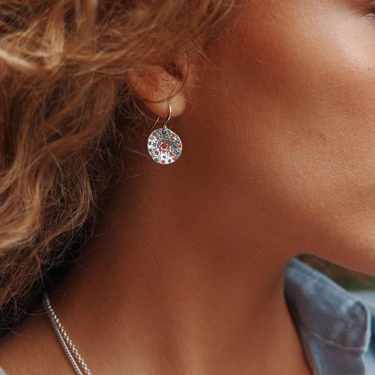 A person wears A&C Oslo Vibrance Silver Coins Earrings with a round, ornate design. Hair partially covers the ear, and a hint of a blue garment suggests the Vibrance Collections vibrant allure.