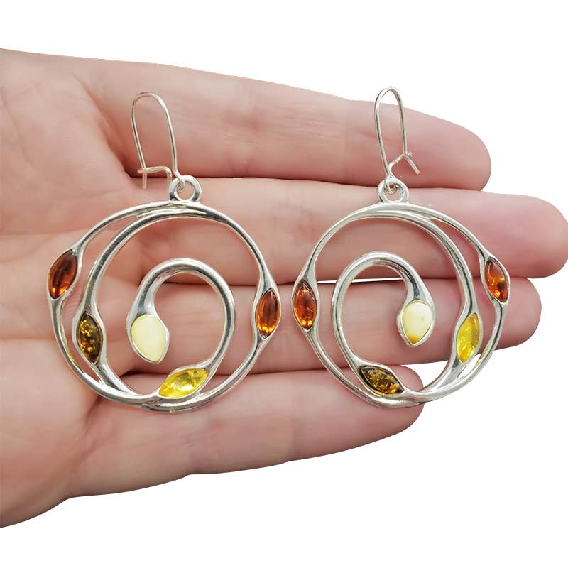 A hand holds a pair of Sterling Silver Earrings on hooks, showcasing curved designs and the captivating beauty of Baltic Amber with multicolored stones in amber, white, and yellow hues.