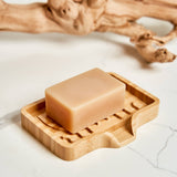 A beige bar of soap on a rectangular bamboo soap lift, with a bare branch placed behind, all set on a white marble surface.