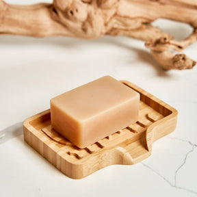 A beige bar of soap on a rectangular bamboo soap lift, with a bare branch placed behind, all set on a white marble surface.