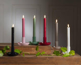 A red Small Pipe Taper Holder (2"H) displays a lit candle on wooden steps beside a green vine, creating modern decor that adds warmth and style to any space.