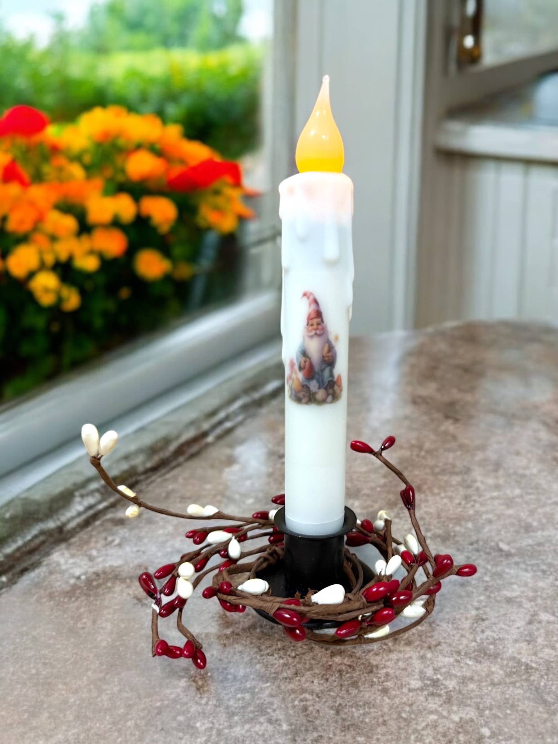 The Spring Gnome Floral Battery-Operated Taper Candle with Timer showcases a charming LED flickering flame, nestled in a red and white berry wreath on the countertop, beautifully complementing the view of spring flowers through the window.