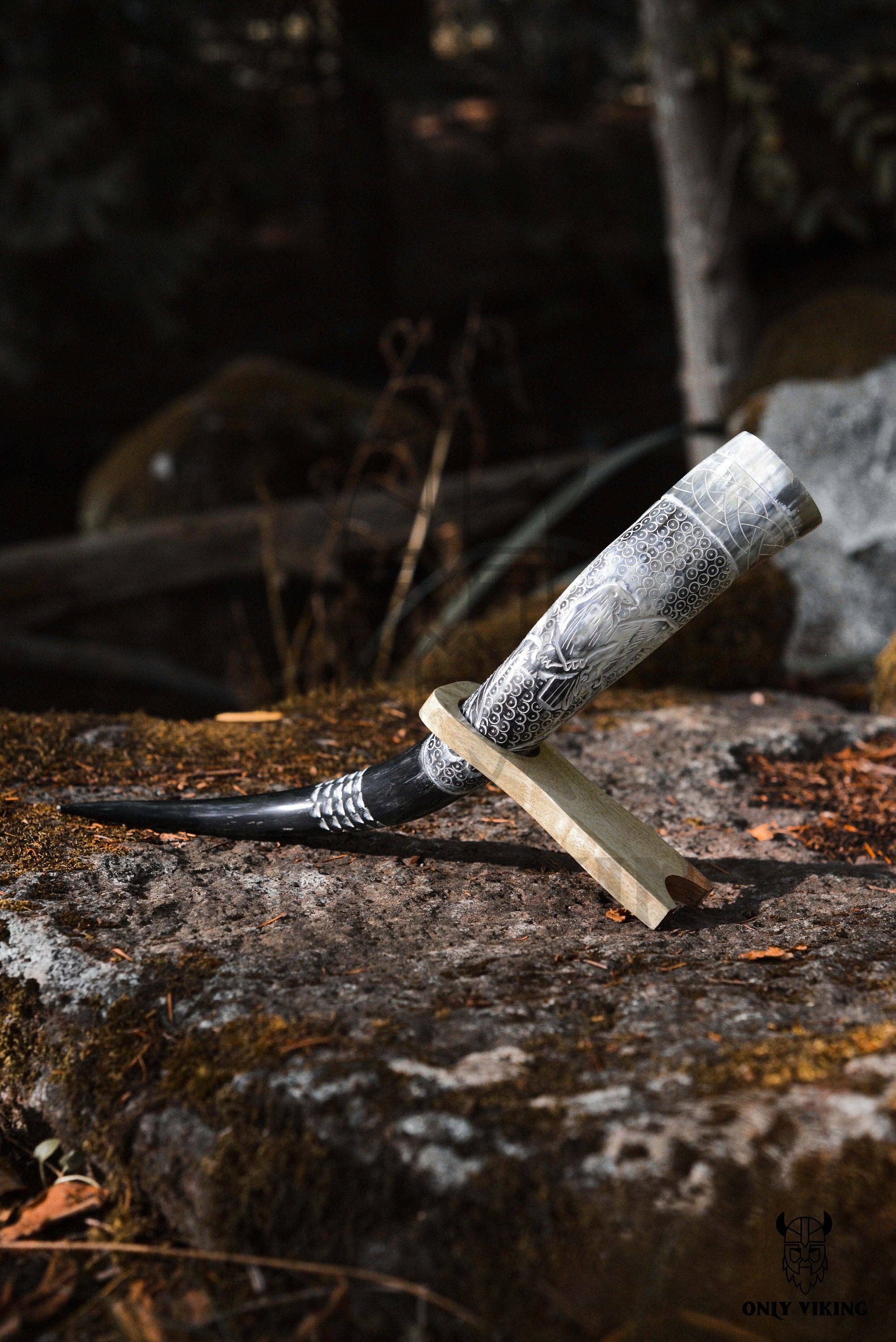 The Viking Drinking Horn, expertly crafted with intricate patterns and supported by a metal stand, is elegantly displayed on a wooden base atop a moss-covered rock outdoors.