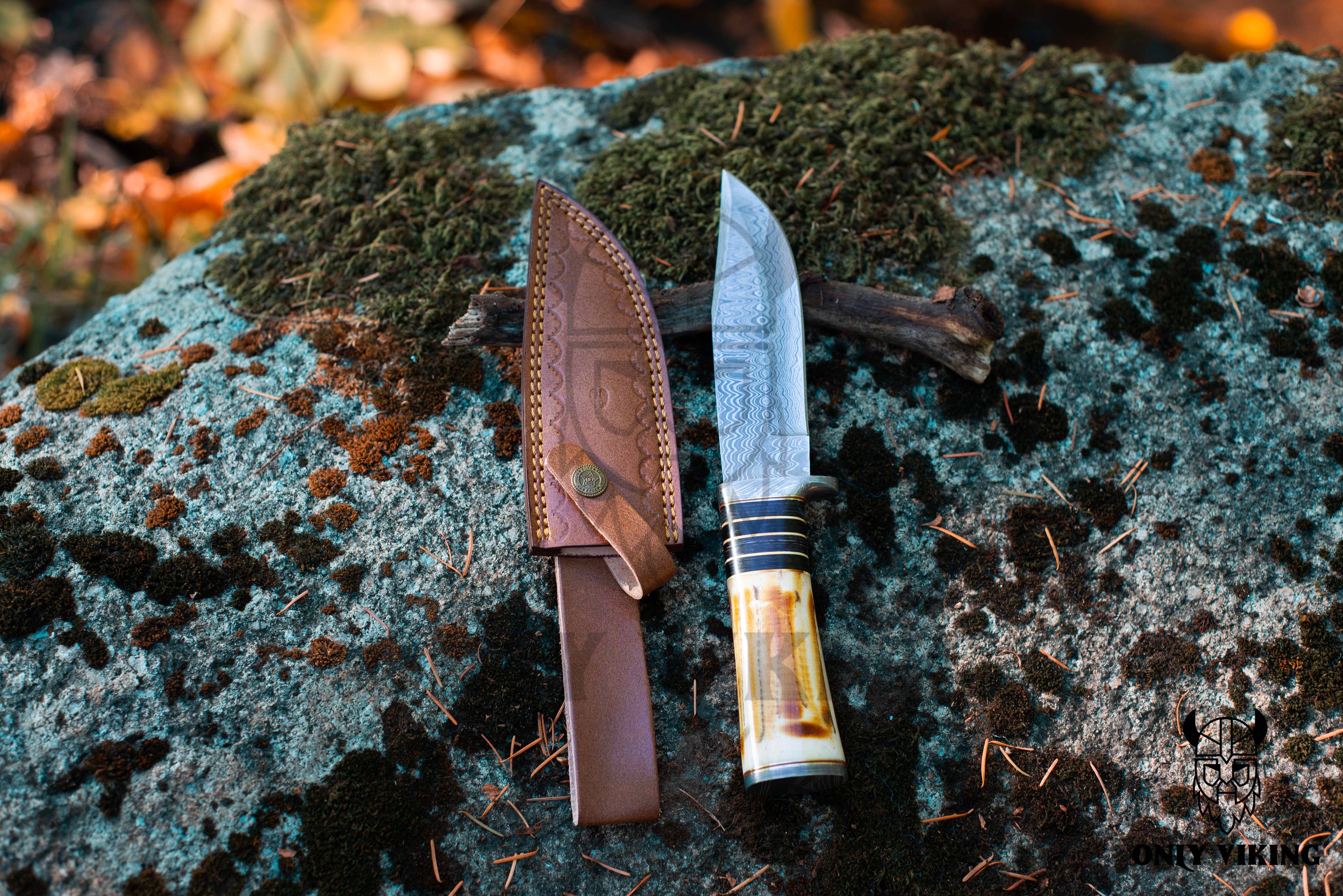 The Damascus Hunting Knife with Locking Leather Sheath showcases its intricate blade on a mossy rock, ideal for outdoor activities.