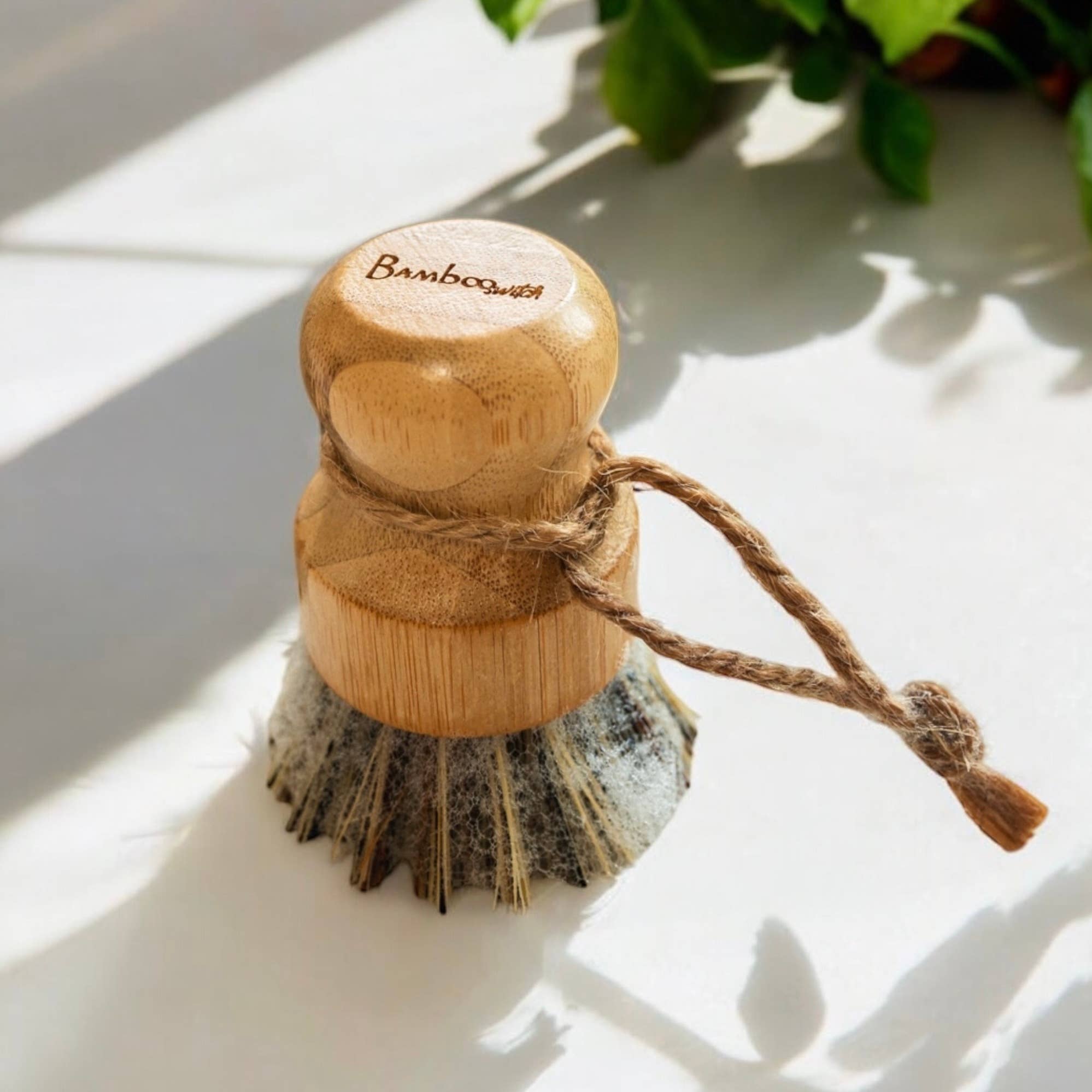 The Scrubber: Bamboo Pot Scruber, with natural bristles and a rope handle, sits on a white surface with sunlight and greenery in the background—an eco-friendly choice for zero-waste cleaning.