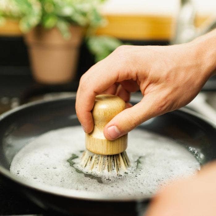 A hand cleans a soapy frying pan using the Scrubber: Bamboo Pot Scruber, an eco-friendly, zero-waste kitchen tool. A blurred potted plant in the background emphasizes this sustainable alternative.