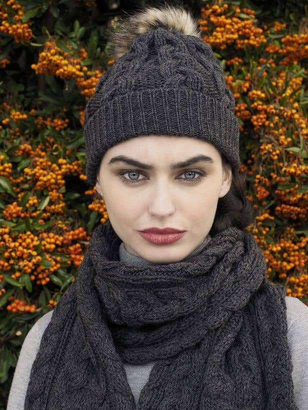 A woman with dark hair wears the Chunky Cable Hat - Charcoal (One Size) as she stands in front of a bush with orange berries.