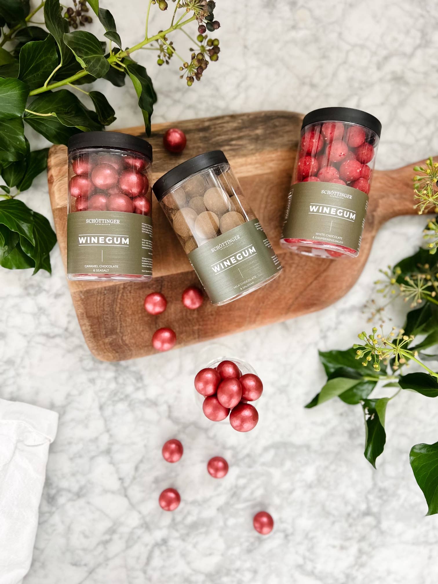 Three jars of Classic Winegum Seasalt & Caramel Chocolate candies are arranged on a wooden board, with scattered red amber candies and surrounded by green leaves on a marble surface.