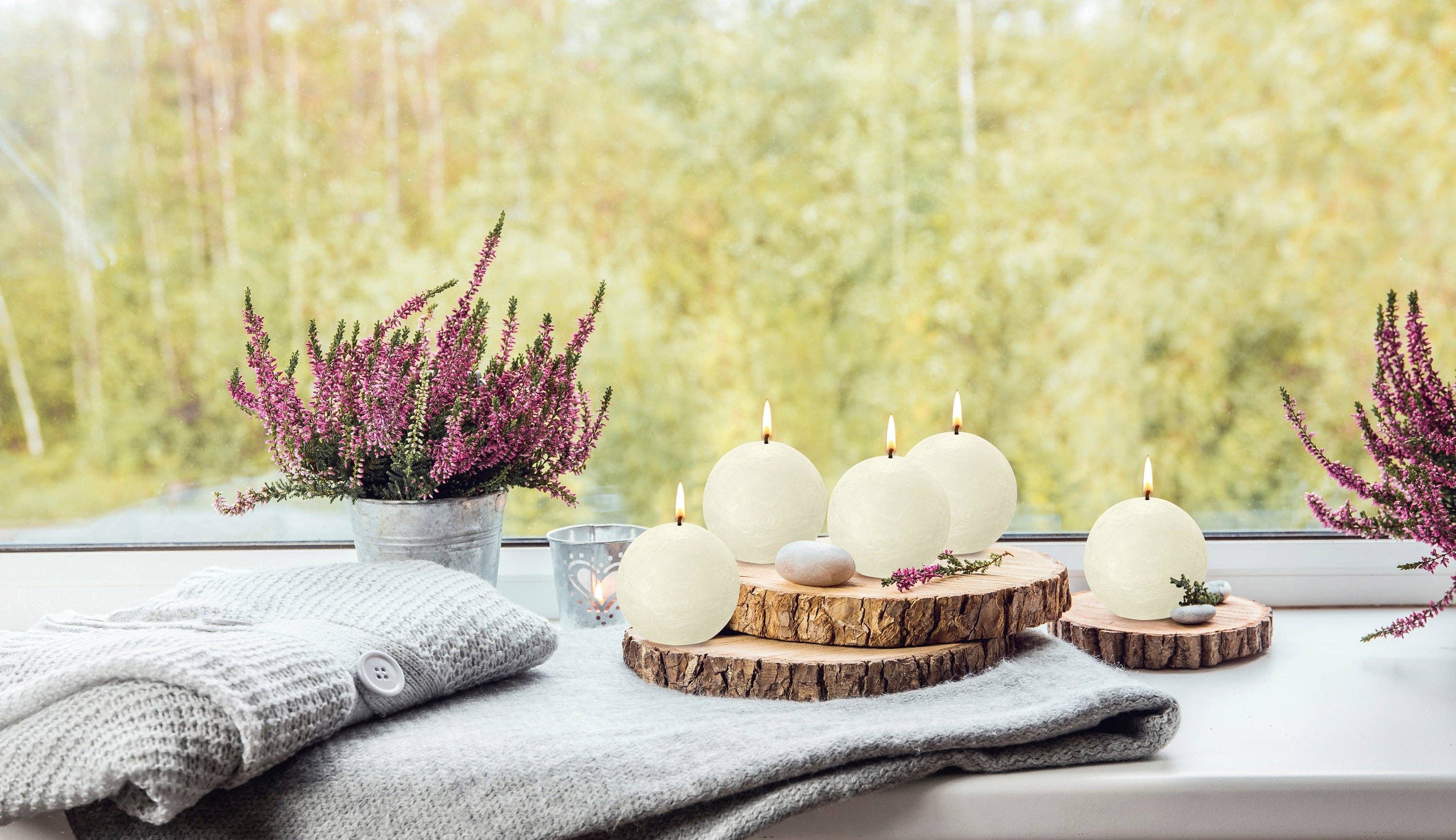 An Ivory Rustic 3" Ball Candle, a folded gray knitted sweater, and potted purple heather flowers create a cozy windowsill arrangement against a softly blurred outdoor backdrop.