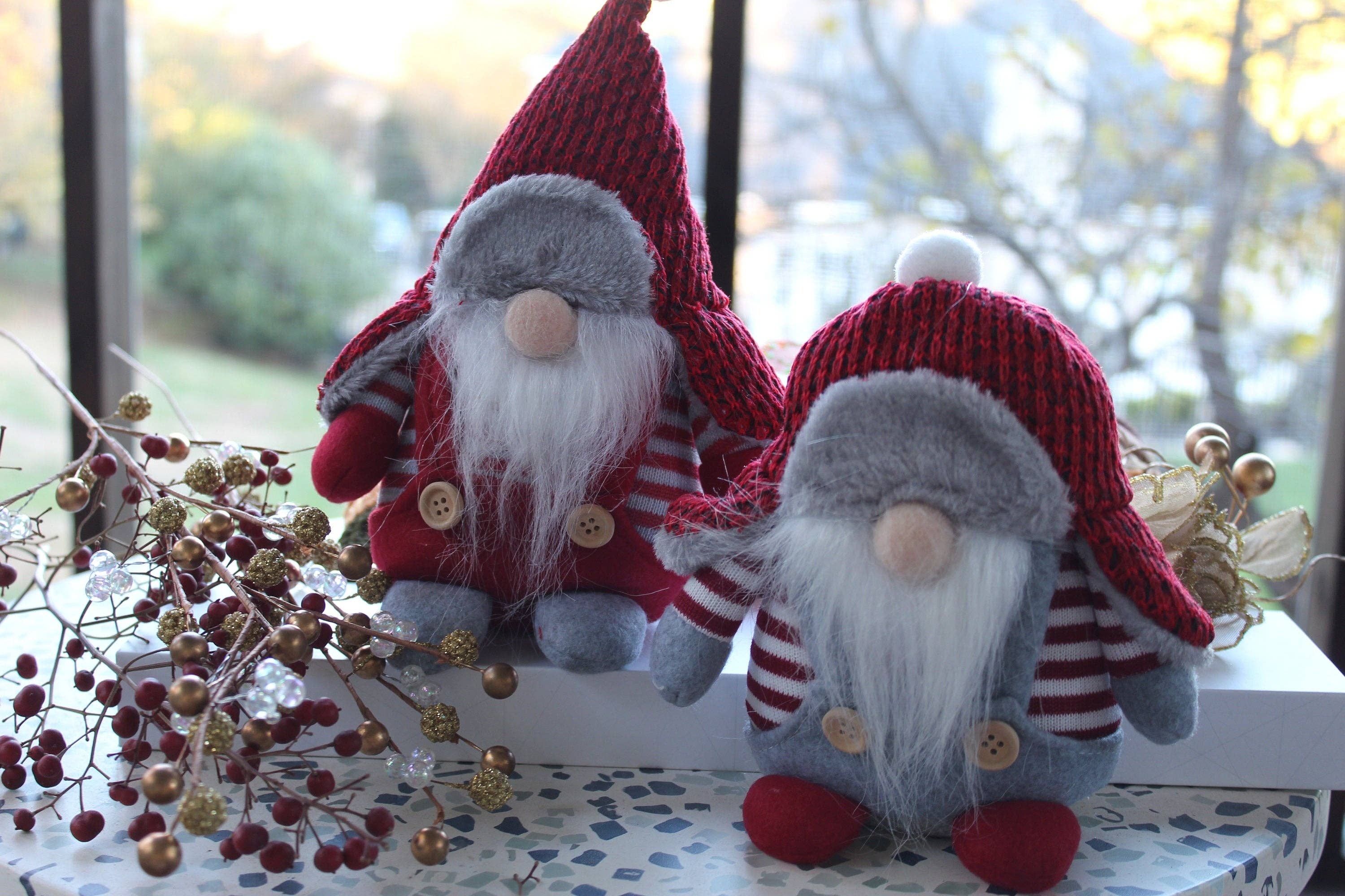 Aldrich and Bernard, two plush gnomes from the Winter Gnomes Christmas collection, inspired by Scandinavian folklore, with red hats and striped shirts, sit on a decorative surface. Branches with red berries are nearby, set against a blurred outdoor background.