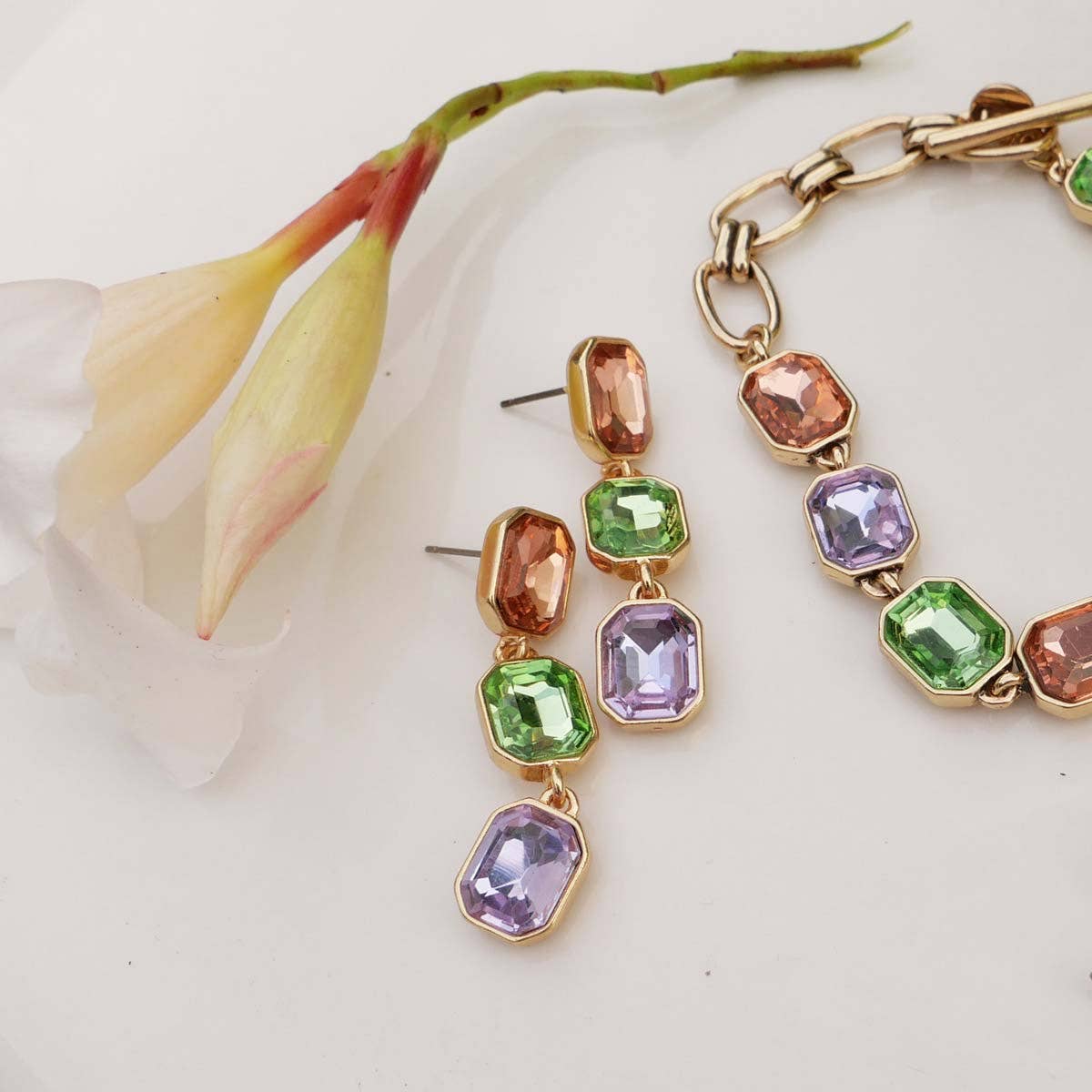 The Multi-Colored Crystal Dangle Earrings, set in elegant gold with pink, green, and purple gemstones, are beautifully displayed next to a pale flower on a white surface, highlighting their exquisite design and complementing a bracelet adorned with emerald cut glass.