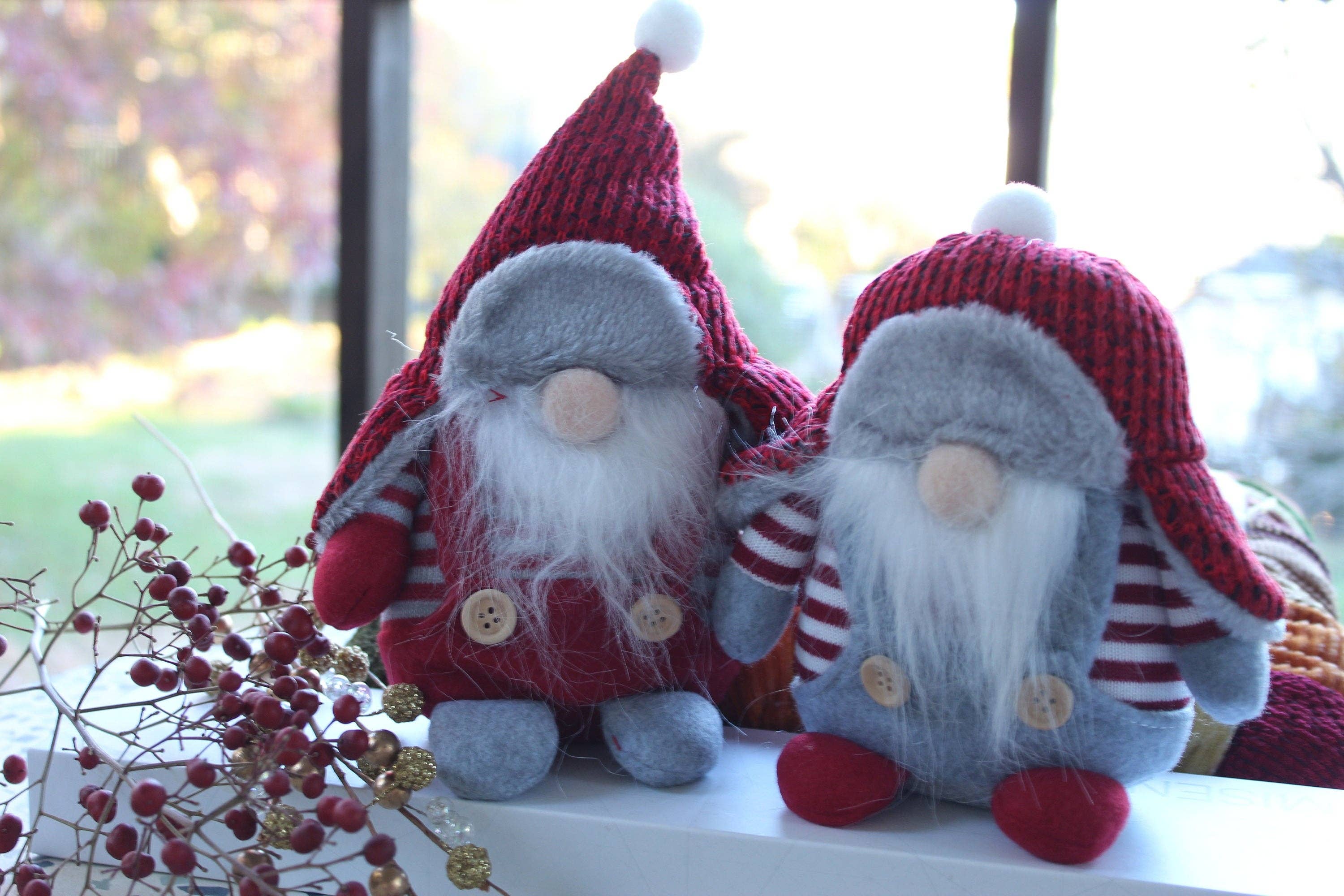 Two plush Winter Gnomes, from the Christmas Gnomes collection and embodying Scandinavian folklore's household spirits, feature gray bodies and white beards. Named Aldrich or Bernard, they are dressed in red hats and outfits, sitting on a surface near a window.