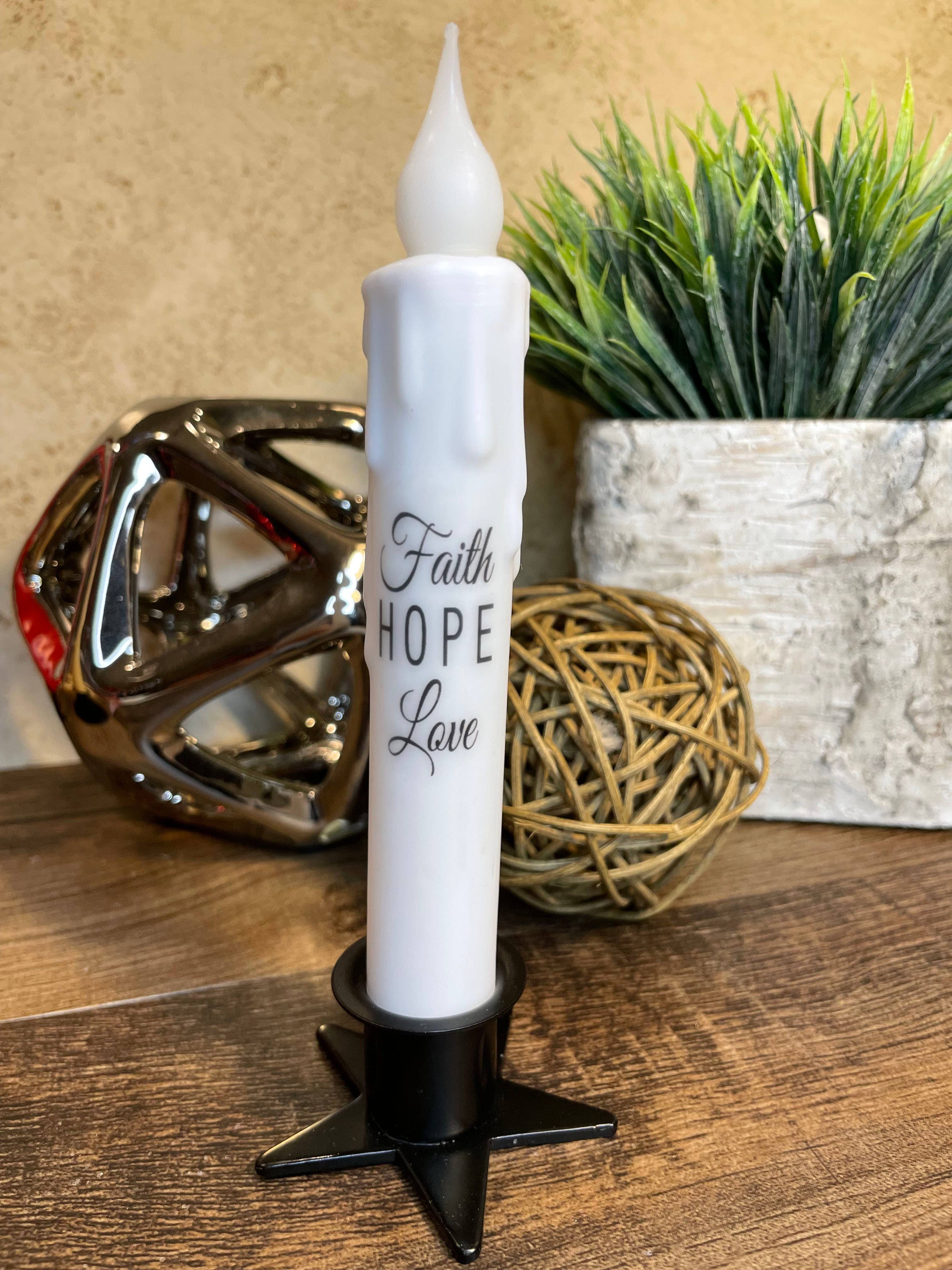 A 7" tapered LED candle with the inscription "Faith, Hope, Love" casts an elegant glow on a wooden surface. This battery-operated timer candle enhances the space beside a metallic sculpture, a wicker ball, and a potted plant.