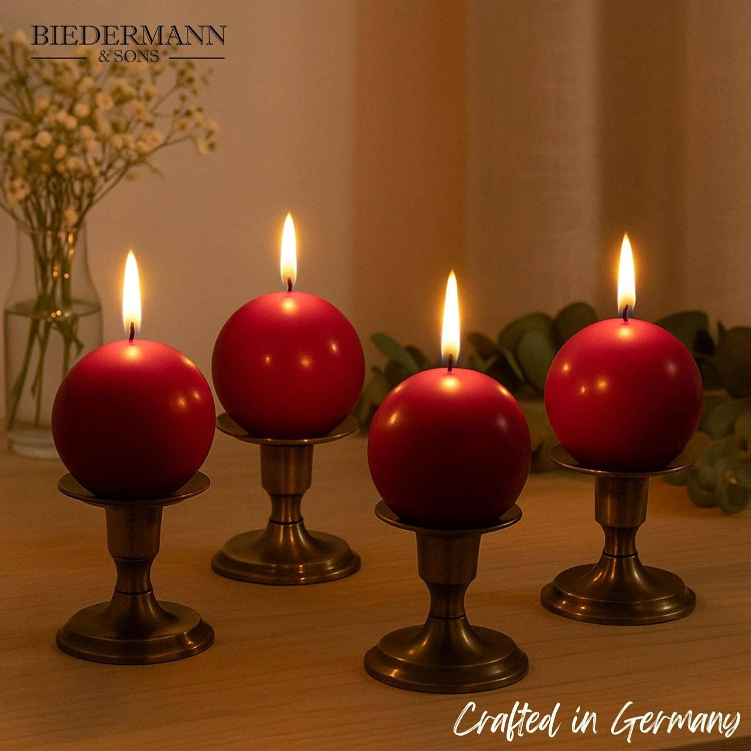 Four lit Burgundy Ball Candles, 2.5" diameter, sit on bronze holders amid flowers and greenery. Text reads Biedermann & Sons and Crafted in Germany, highlighting these elegant decorative candles.