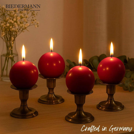 Four lit Burgundy Ball Candles, 2.5" diameter, sit on bronze holders amid flowers and greenery. Text reads Biedermann & Sons and Crafted in Germany, highlighting these elegant decorative candles.