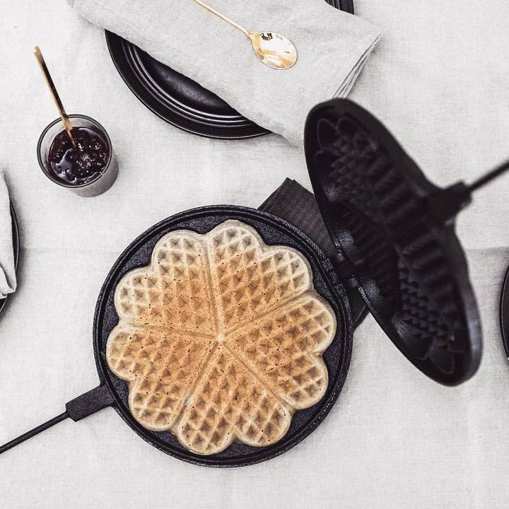 The Skeppshult Waffle Iron 8.25 (21 cm) sits on a table with a heart-shaped waffle, a plate, gray napkin, gold spoon, and a small jar of jam.