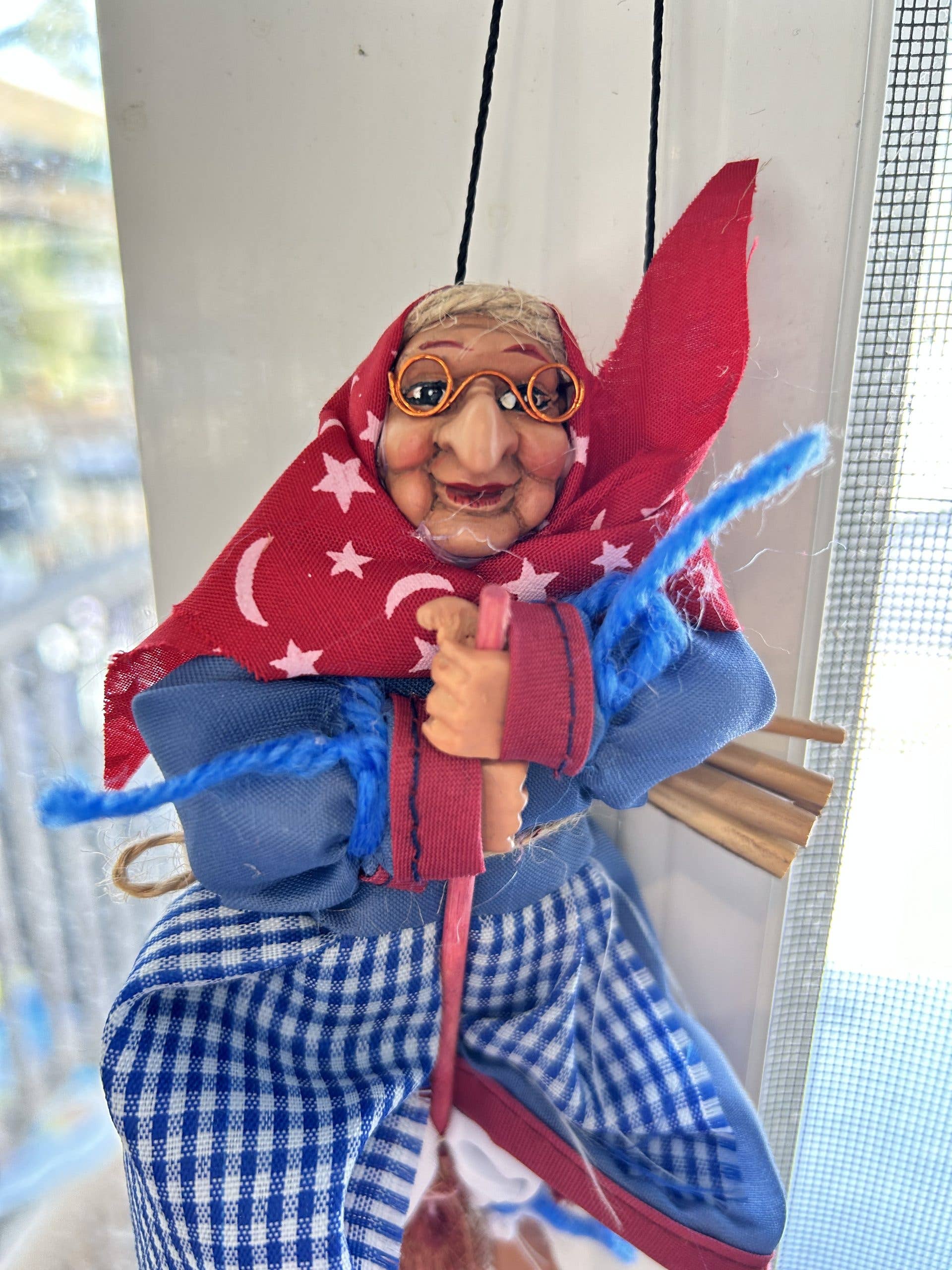 The Opalina Blue Kitchen Witch Figurine, 7" tall, features glasses, a red scarf with stars and moons, blue shirt, checkered skirt, and holds a broom—perfect as a good luck charm to hang in your kitchen window.