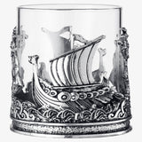 The 12 OZ Viking & Mythological Whiskey Drinking Glass features a clear design with ornate silver ship and wave motifs wrapping elegantly around its base and sides.