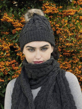 A woman wears the Chunky Cable Hat - Charcoal (One Size), a merino wool pom-pom hat, and matching scarf while standing by a bush with clusters of orange berries.