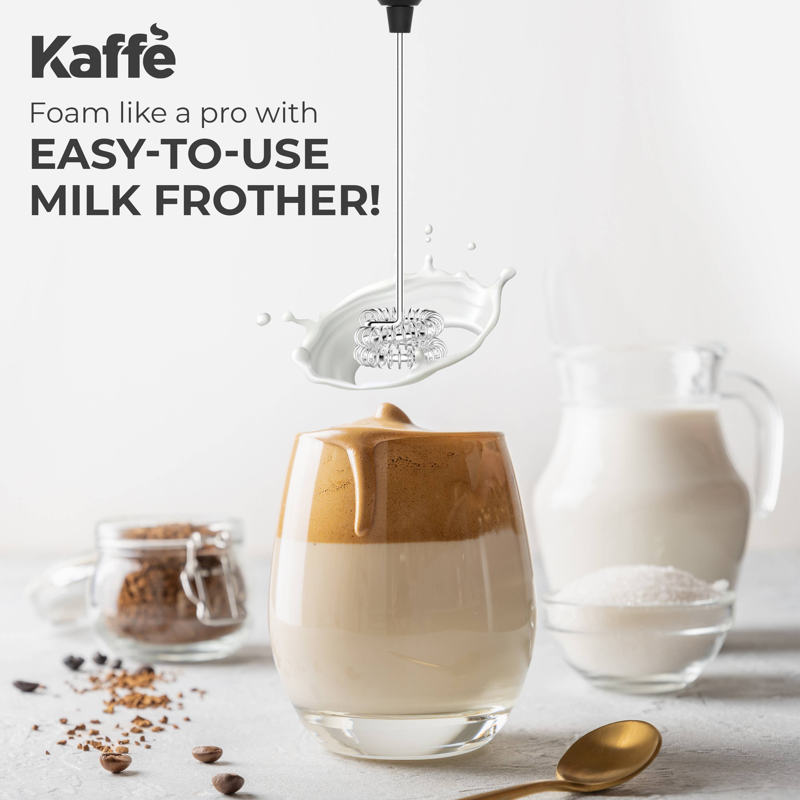 The Kaffe Handheld Milk Frother with Stand (USB Rechargeable, Stainless Steel) whips milk above a glass of layered coffee, surrounded by cocoa, milk, sugar jars, coffee beans, and a spoon.