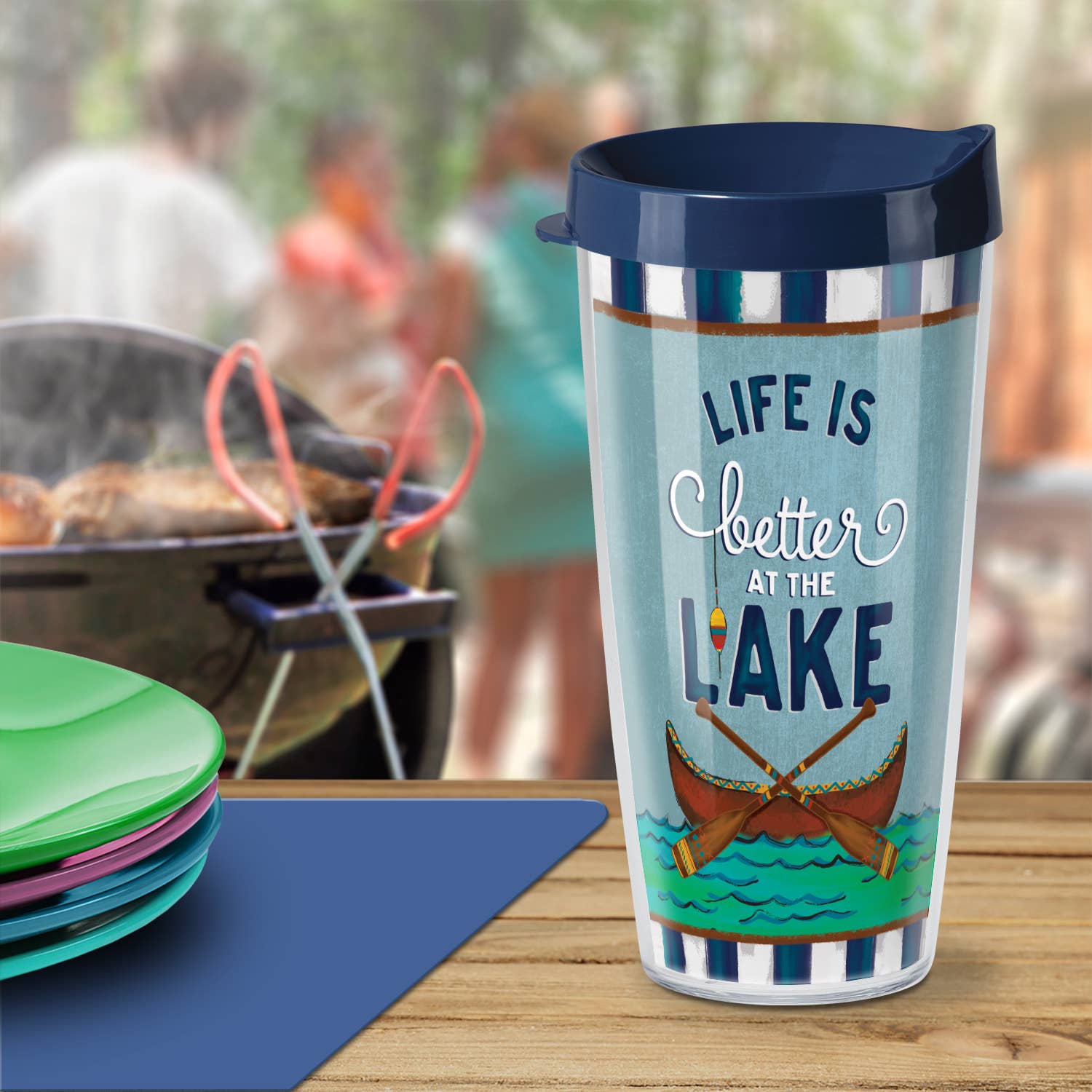 The "Life is Better at the Lake" 22 oz. Double Wall Insulated Tumbler is dishwasher-safe and adorned with the text and an illustration of canoes, featuring a backdrop of artistically blurred people and a barbecue to encapsulate the essence of lakeside relaxation.