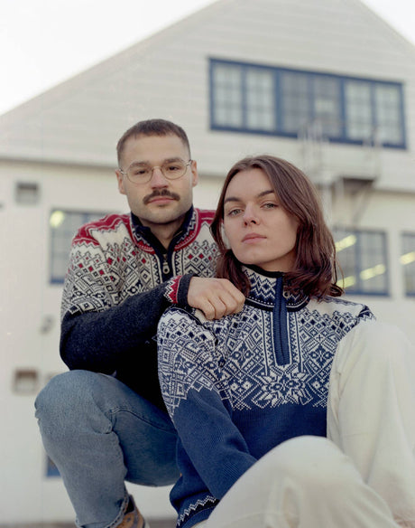 A man and a woman wearing Norlender Knitwear – Narvik Ski Sweater (312) in charcoal pose in front of a light-colored building with blue-trimmed windows.