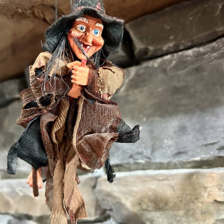 A close-up of the Walburga Brown Kitchen Witch Doll shows its exaggerated features and ragged clothes before a stone wall. Traditionally handmade in Germany, kitchen witch dolls are good luck charms for the home.