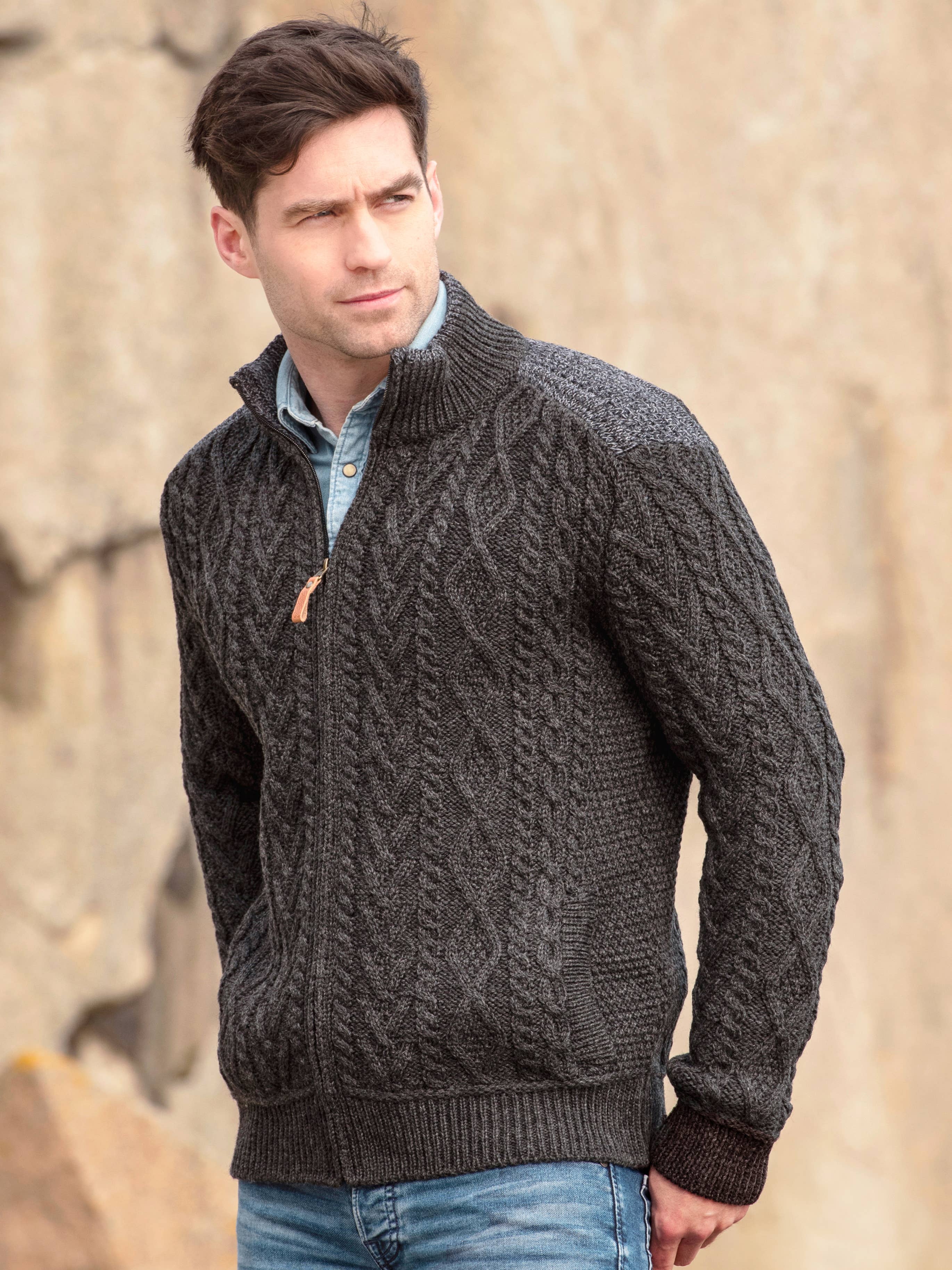 A man wearing a Mans Zip 100% Merino Wool Cardigan in Charcoal over a button-up shirt stands outdoors against a blurred rocky background.