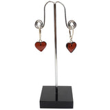 Heart Cherry Amber Sterling Silver Earrings with red heart-shaped pendants, reminiscent of cherry amber, are displayed on a black upright stand.