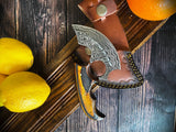 The Alaskan Ulu Style Blade, a high carbon Damascus steel knife with ergonomic design, wooden handle, and leather sheath, is shown on a wooden surface beside lemons and an orange.