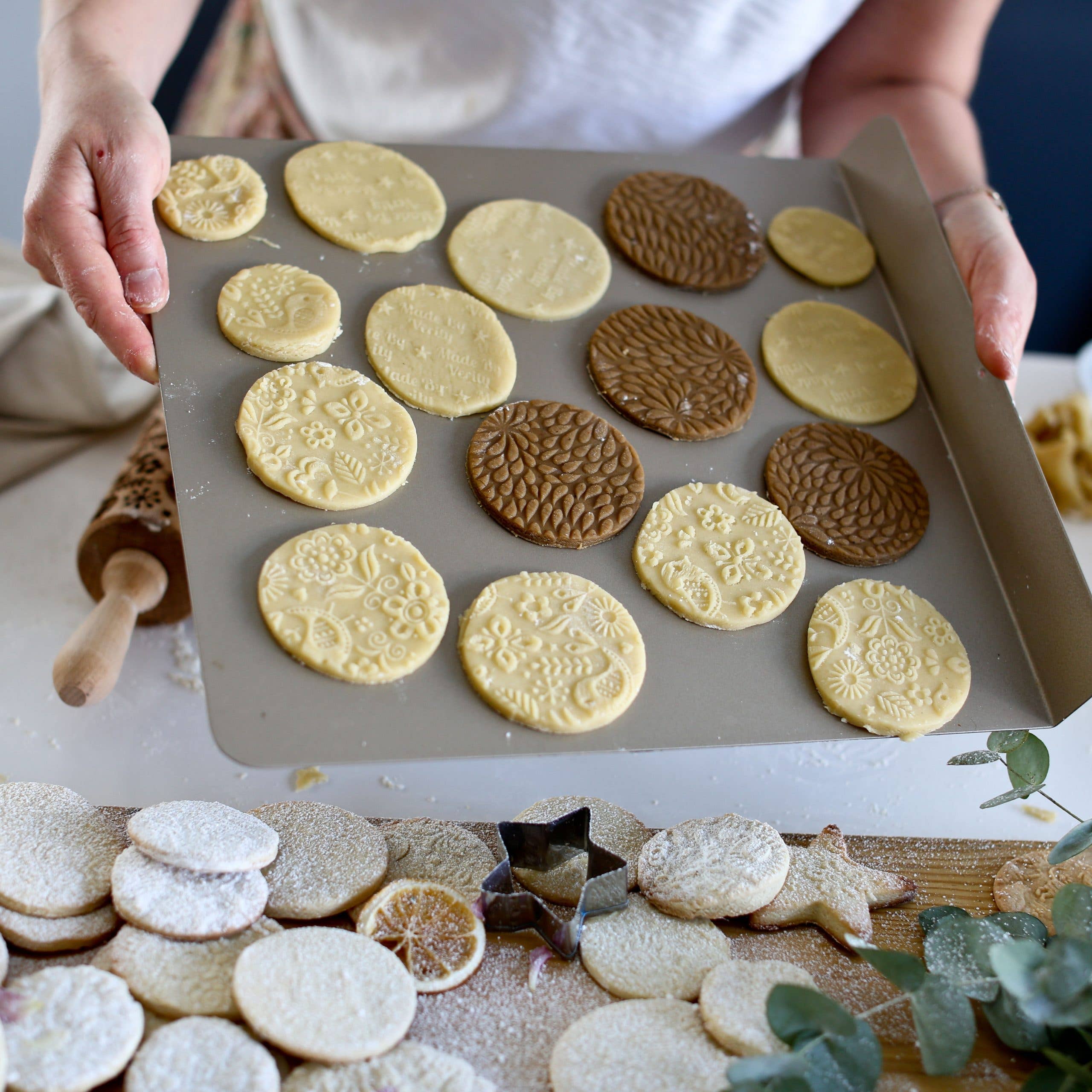 A person holds a tray of unbaked stamped plain and chocolate cookies with more cookies on the table. An Autumn Leaves Embossing Rolling Pin is ready to imprint its intricate designs on the next batch.