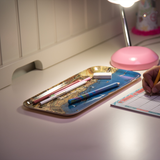 A hand writes on paper next to a birchwood tray holding pens and erasers, beneath a pink desk lamp on a white desk.
