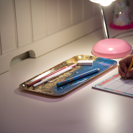 A hand writes on paper next to a birchwood tray holding pens and erasers, beneath a pink desk lamp on a white desk.