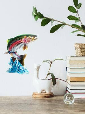 Jumping Trout Wall Art decor featuring a vibrant trout leaping from the water, positioned alongside a book stack and a plant in a llama-shaped pot on the shelf.