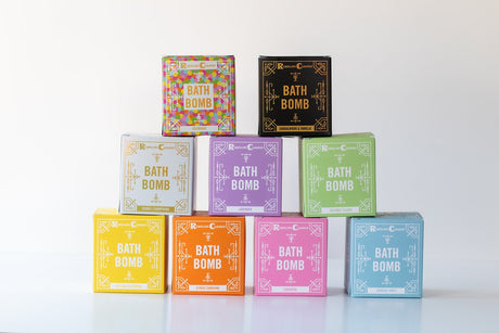 Nine colorful bath bomb boxes, including the Soap: Sandalwood & Vanilla Bath Bomb, are arranged in a pyramid on a white surface, each featuring unique scents and eye-catching designs.