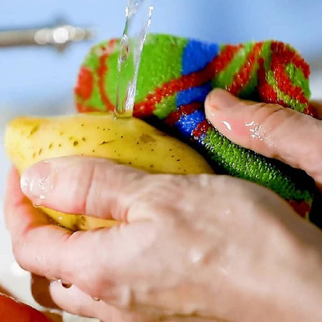 A person scrubs a yellow potato under running water using the Scrubbie: Skrubba XL Scrub Cloth for scratch-free cleaning.