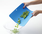 Hands pour chopped celery from a flexible, BPA-free cutting board into a clear glass bowl. Only the hands, cutting board, celery pieces, and bowl are visible.