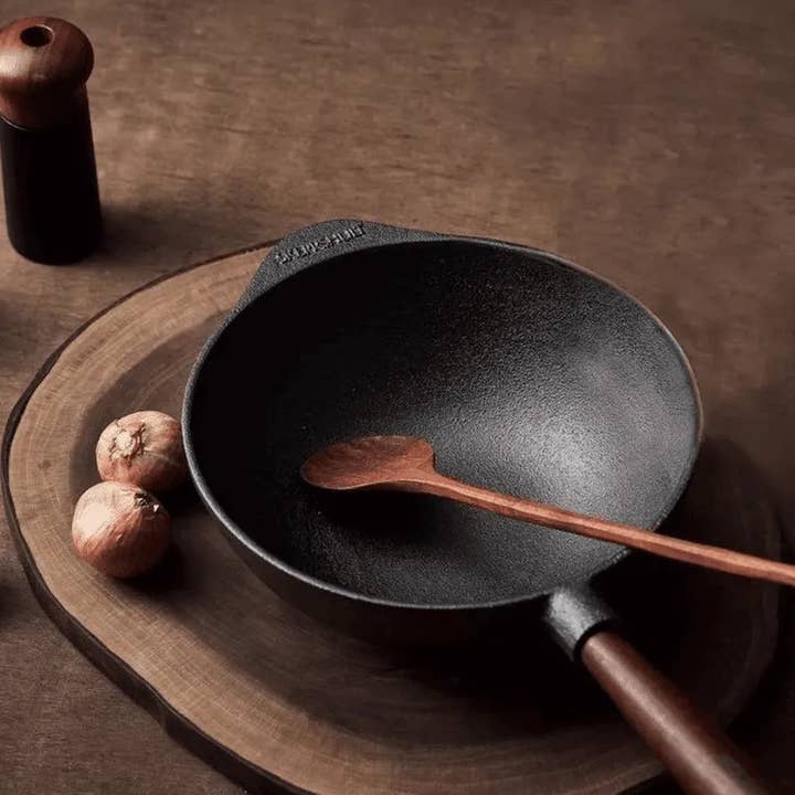 A Skeppshult Wok With Walnut Handle 12.2 sits on a wooden board with a wooden spoon, two onions, and a pepper grinder. Its natural non-stick surface is highlighted in this elegant kitchen display.