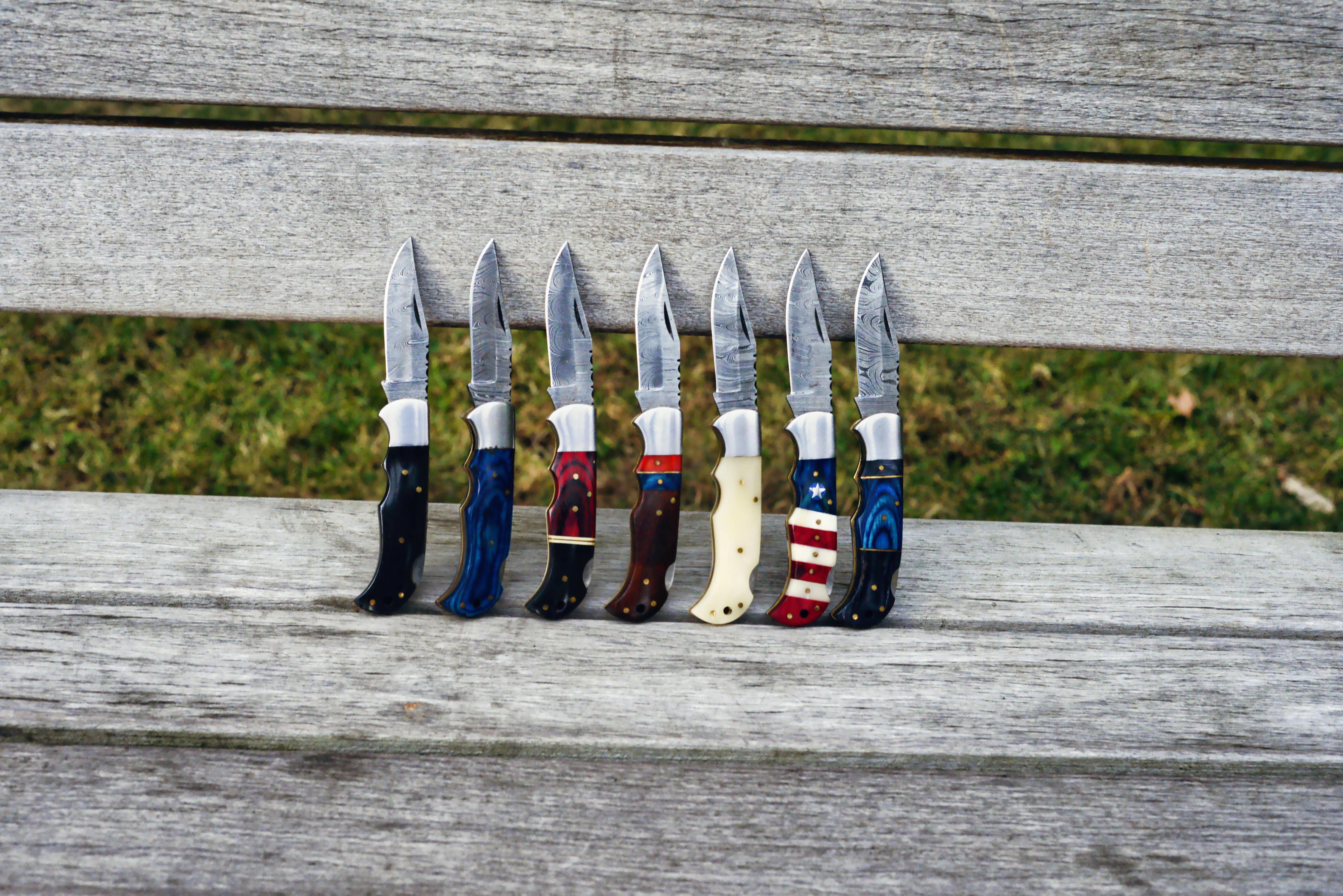 Displayed outside on a wooden bench seat are seven handcrafted pocket knives, each with unique handle designs, including the striking Camel Bone Damascus Folding Knife. These exquisite tools blend artistry with functionality, making them perfect as personalized gifts.