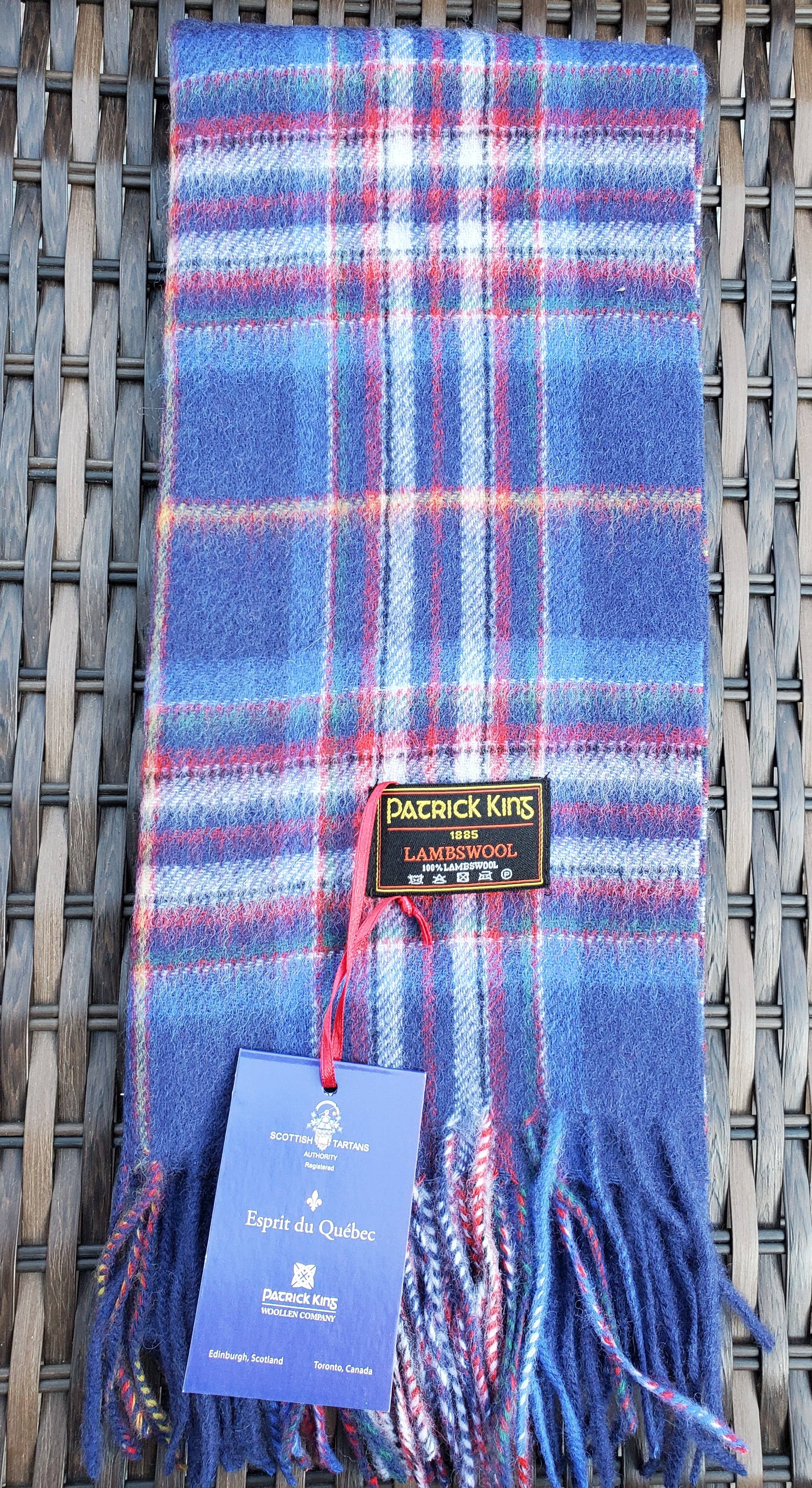 The Esprit du Quebec Lambswool Scarf in blue and red plaid, featuring fringed ends, is shown on a woven surface. The Patrick King Lambswool label and an attached blue authenticity tag are visible.