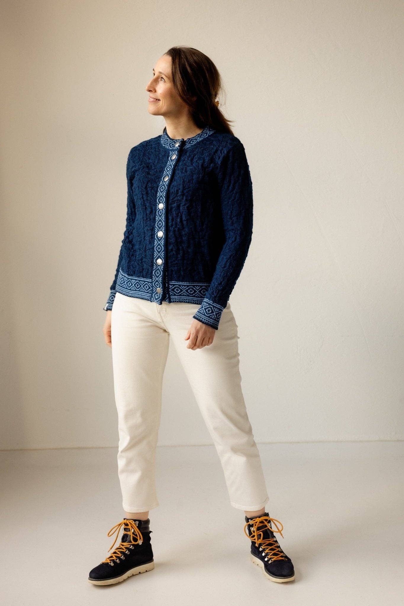 Indoors, a person wears the Bryggen Women's Norwegian Cardigan (White) with white pants and black boots with orange laces, looking slightly to the side.