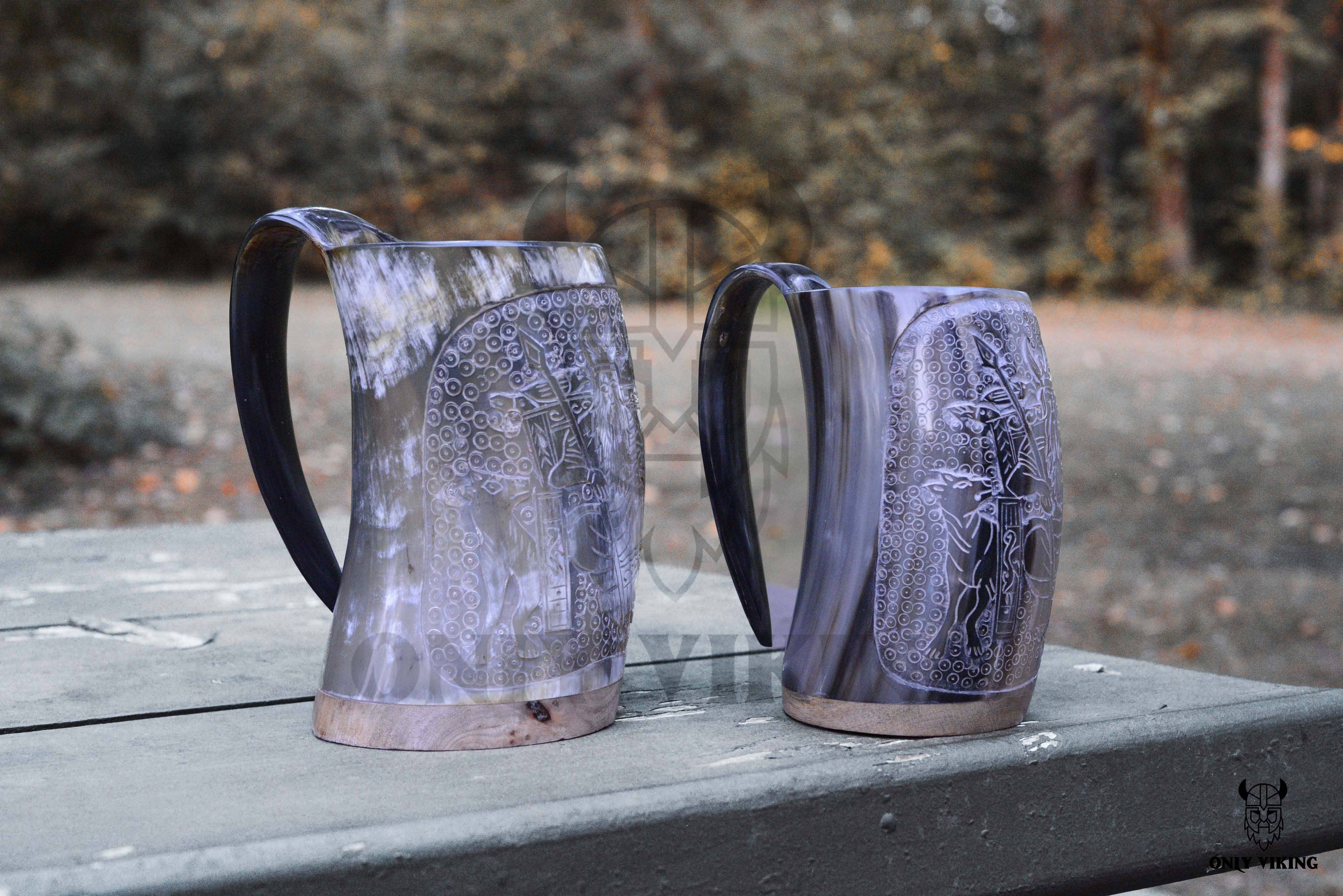 Two ornately designed, hand-carved "Horn: Viking Tankard" drinking mugs with handles rest on a wooden picnic table outdoors, set against a blurred natural background.