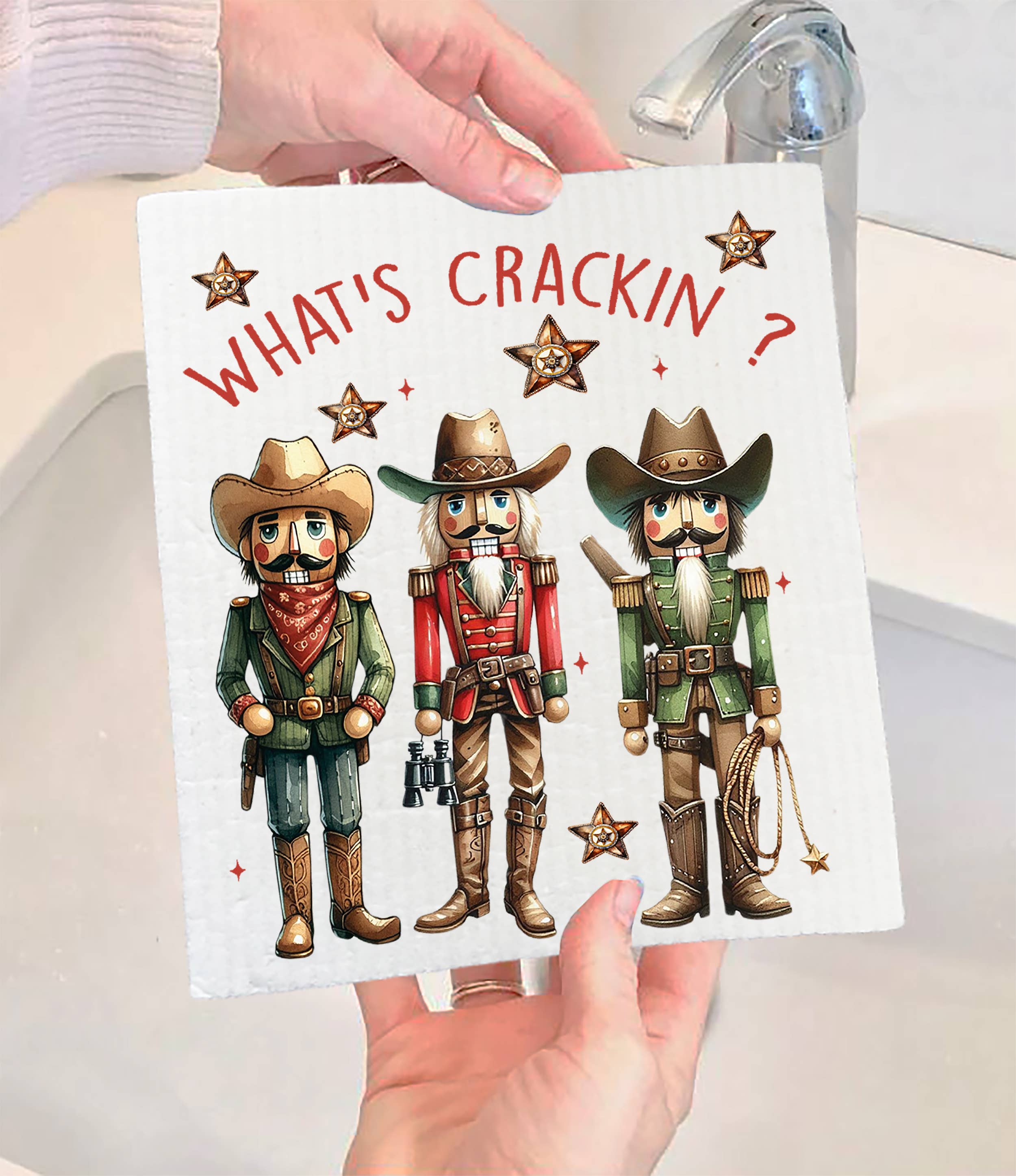 Hands holding a Swedish dishcloth named "Christmas Western Cowboy Nutcrackers," adorned with three cowboy nutcracker figures and the bold text "What's Crackin?" This dishcloth is both biodegradable and stylish for your kitchen.