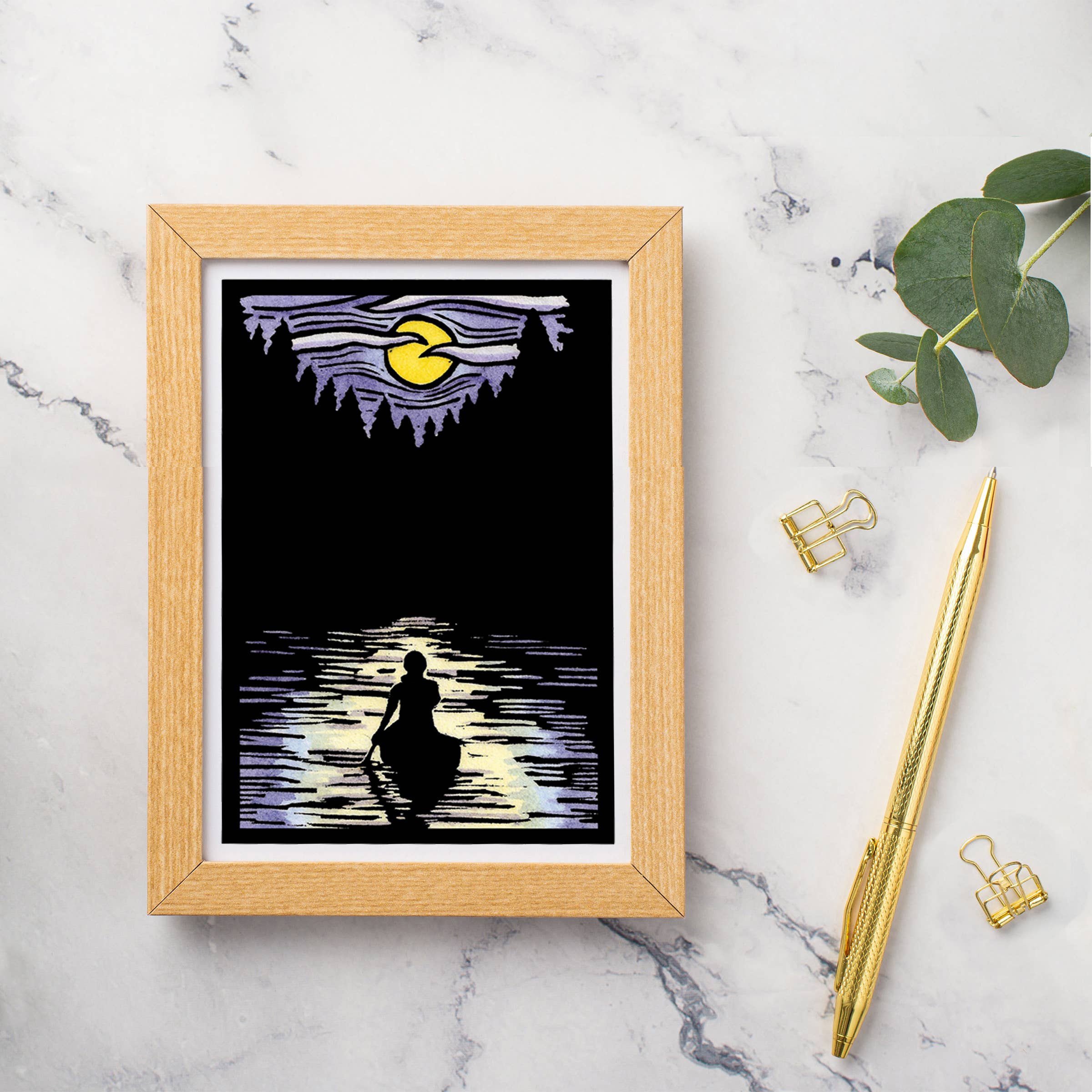 Framed artwork titled "Solitude Canoe" by Sarah Angst, featuring a person in a boat on mirrored waters under a crescent moon and purple sky. This captivating piece, echoing the style of linocut prints, is displayed on a marble surface alongside matching greeting cards, paper clips, and eucalyptus leaves.